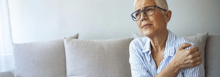 Person seated on a sofa holding the shoulder, illustrating rotator cuff injury or chronic shoulder discomfort.